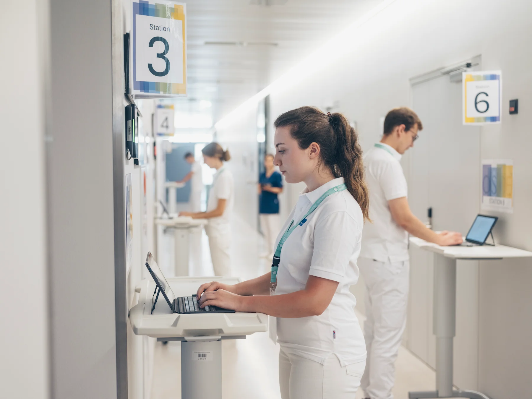 Healthcare professionals in white uniforms working on laptops in hospital corridor, managing patient records and clinical documentation.