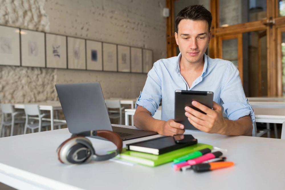 A young man in a light blue shirt sitting at a white desk in a modern workspace, using a tablet with a laptop, headphones, and colorful stationery nearby.