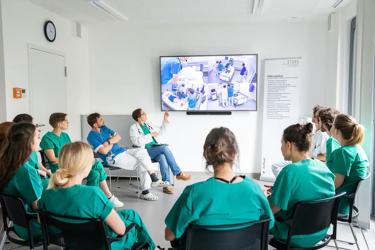 Medical students in scrubs watching multi-angle simulation training on screen with instructor, STUPS banner highlighting crisis resource management.
