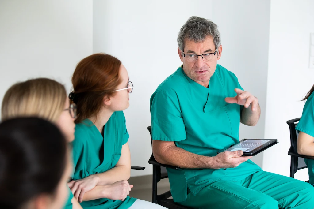 SIMStation Medical team in green scrubs collaborating during training, leader with tablet guiding discussion.