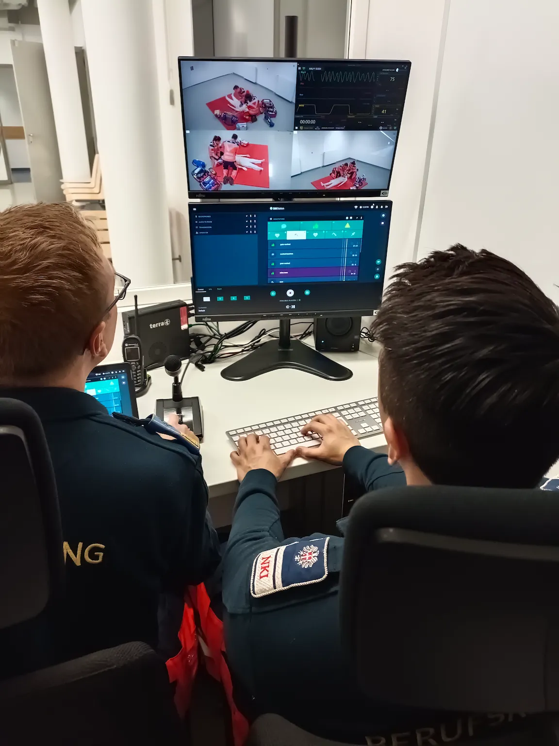 Emergency training personnel at workstation monitoring medical simulation feeds and vital signs on dual screens.