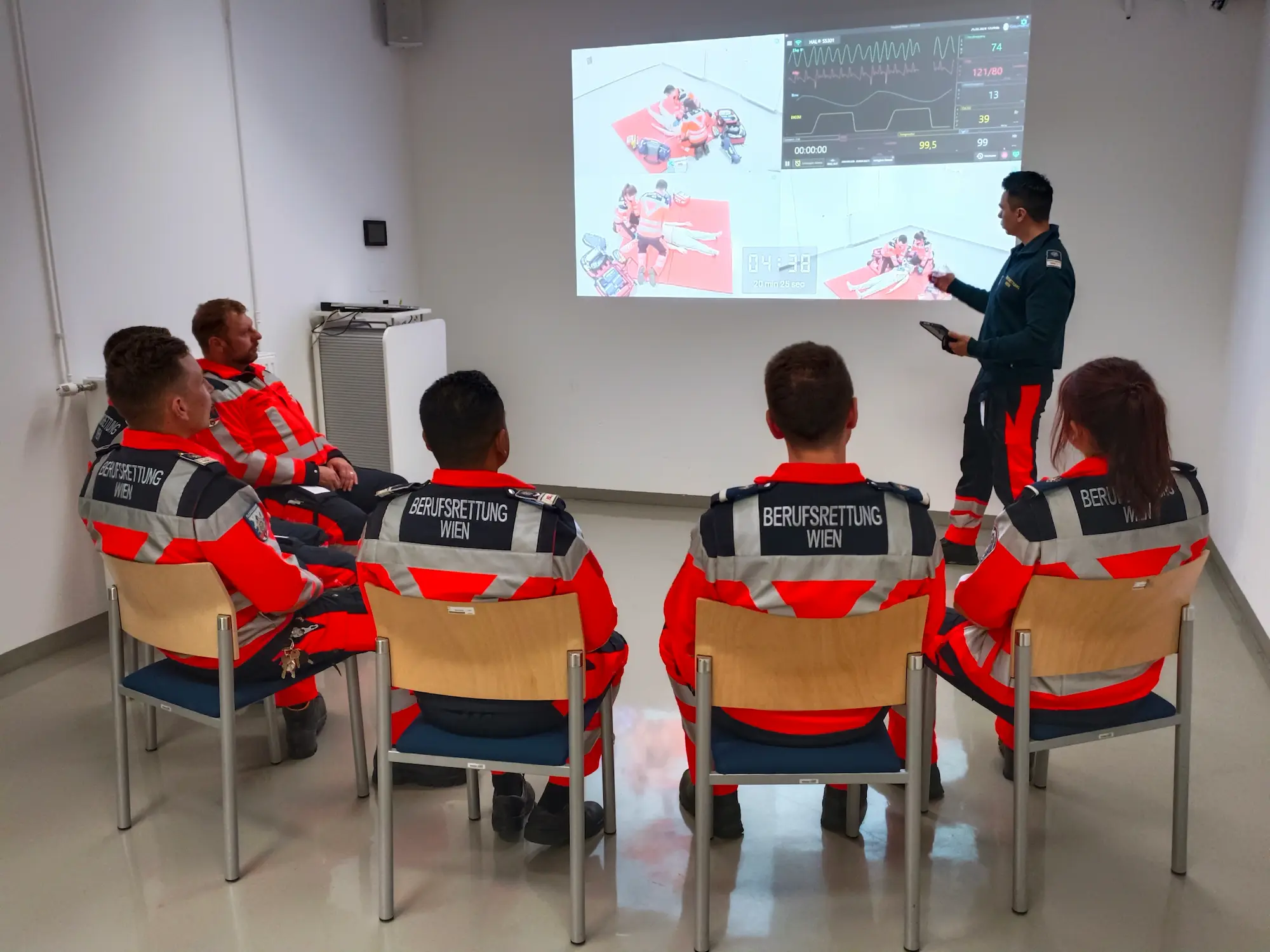 Professional rescue workers from Vienna attending medical simulation briefing with projected vital signs and scenario data.
