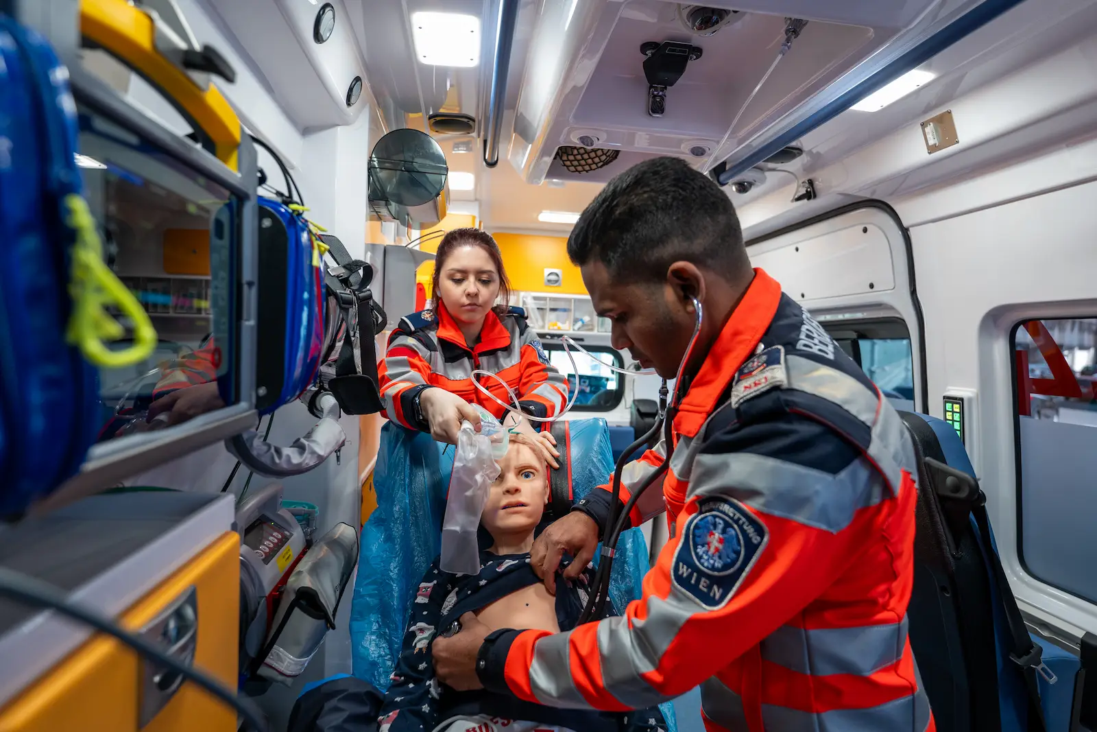 Two paramedics from Berufsrettung Wien inside a fully equipped ambulance performing a clinical assessment and respiratory support on a child-sized patient manikin during a high-fidelity emergency simulation.