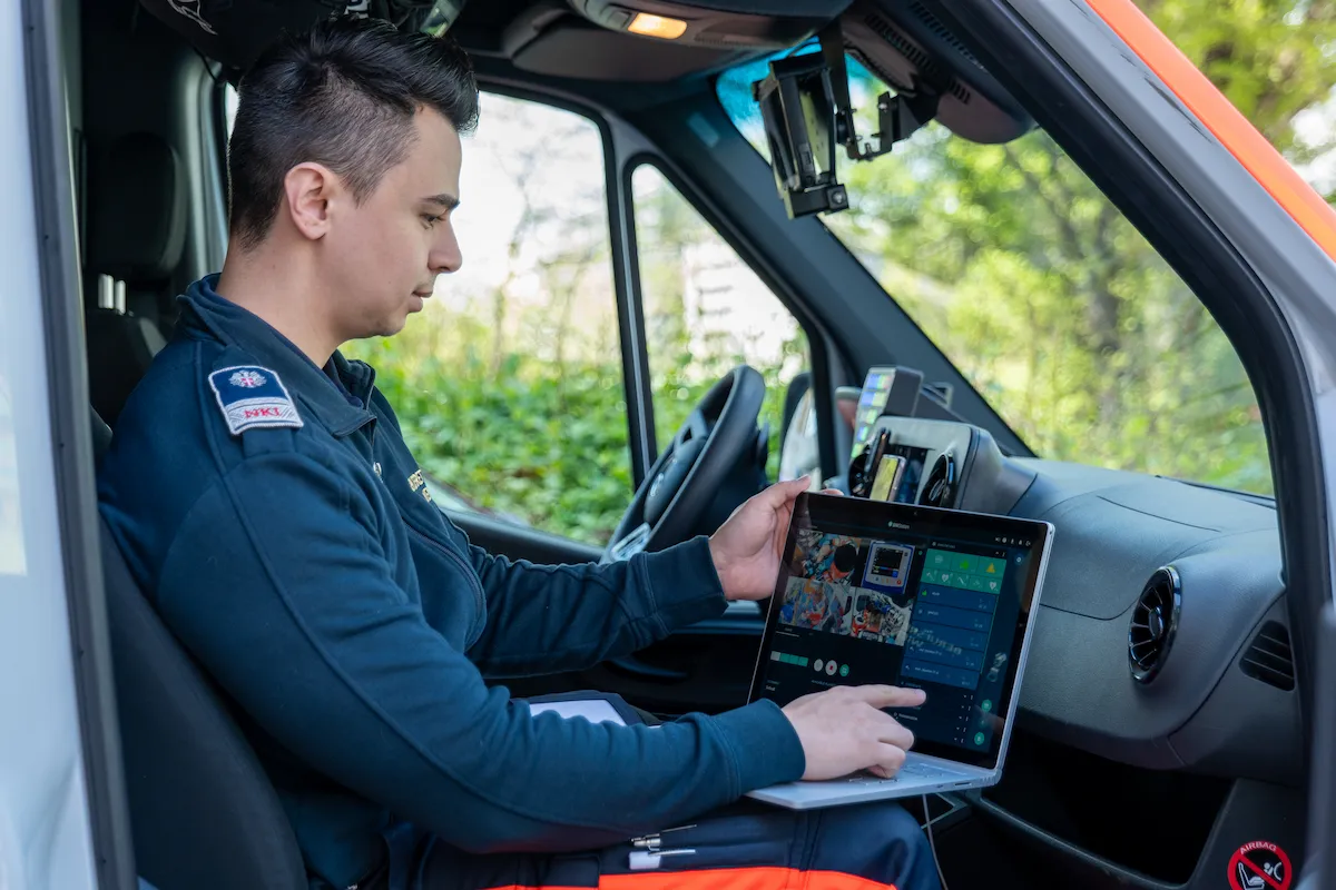 An emergency medical professional from Berufsrettung Wien sitting in an ambulance cab using a laptop to review multi-camera SIMStation simulation footage for mobile field training debriefing.