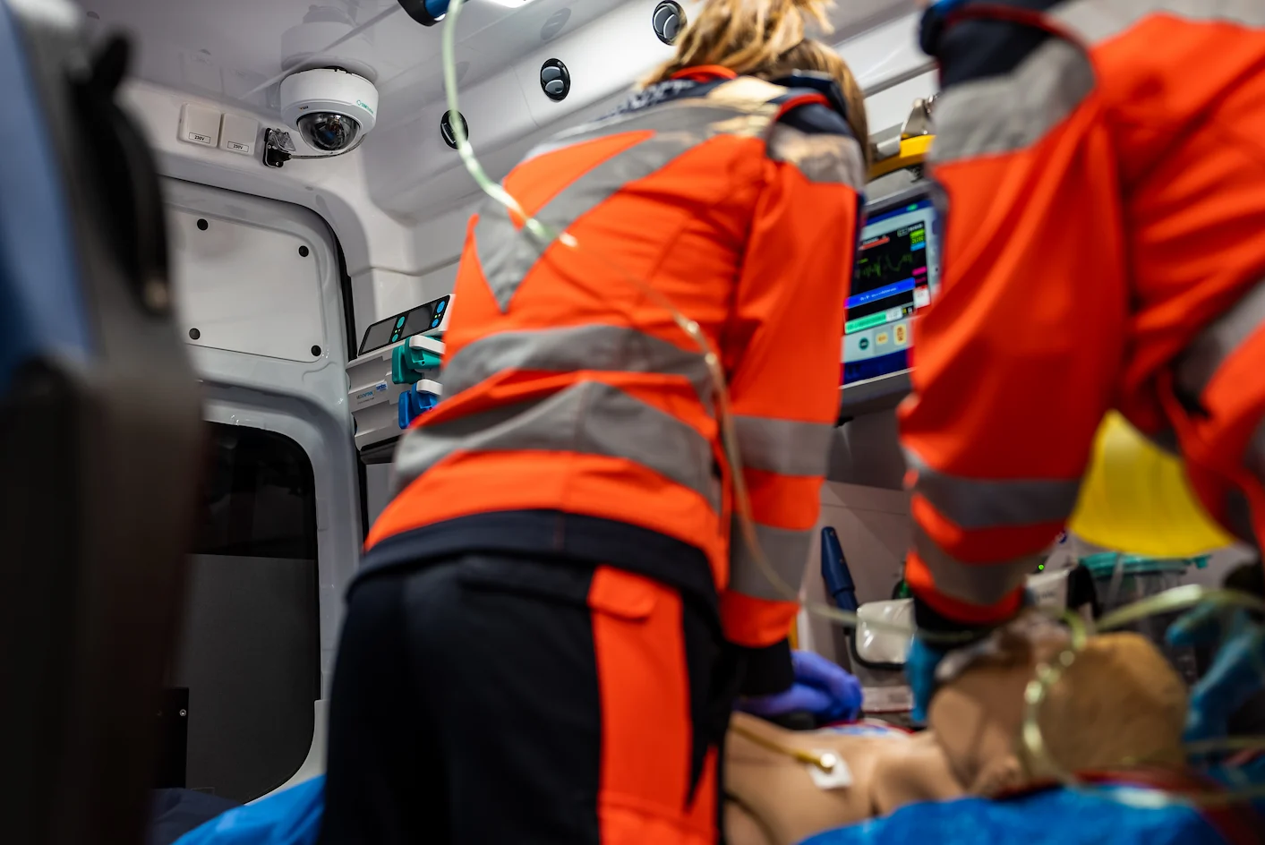 Interior view of an ambulance where a Berufsrettung Wien emergency team performs life-saving procedures on a manikin, monitored by a ceiling-mounted SIMStation camera for high-fidelity simulation recording.