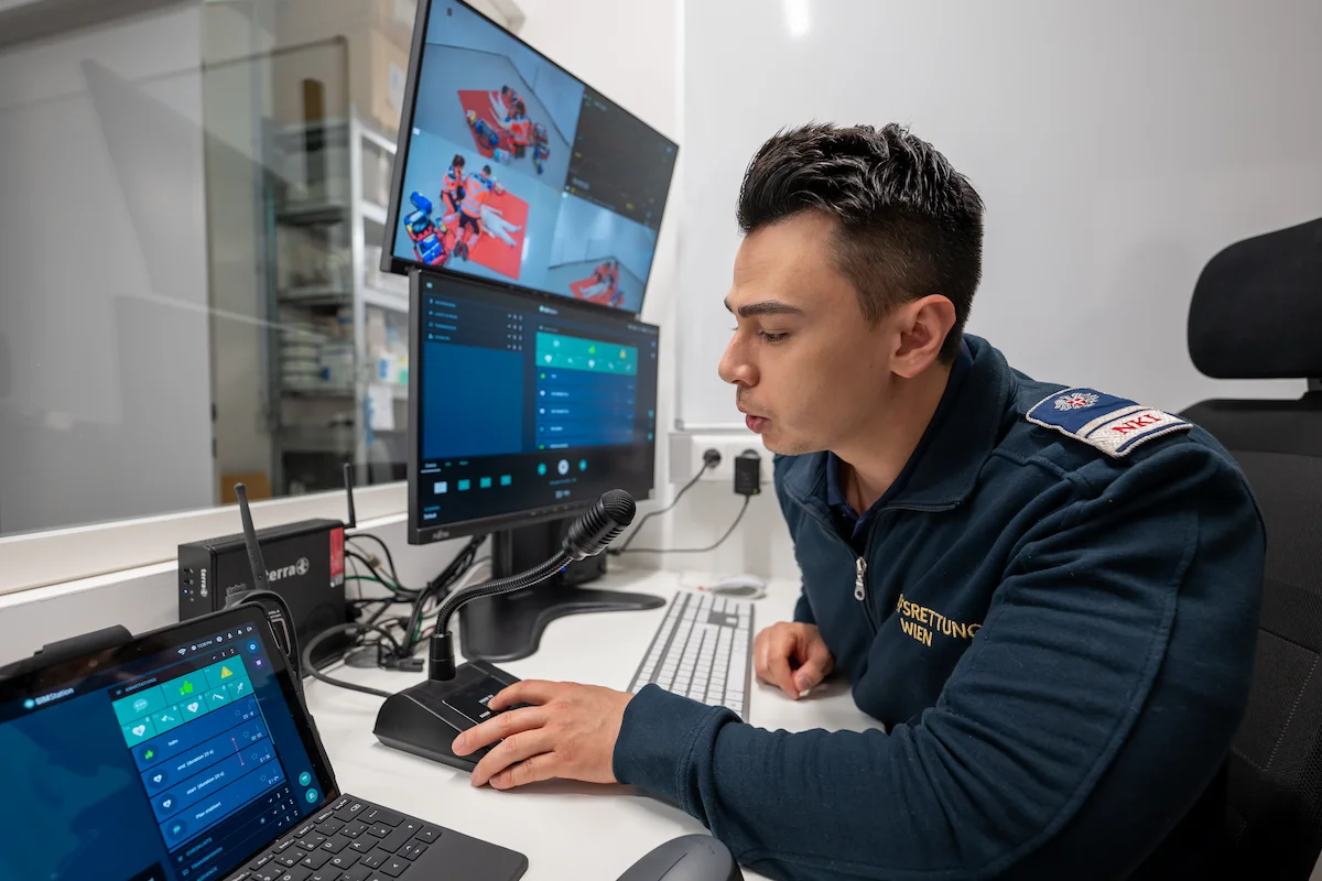 An instructor from Berufsrettung Wien at a SIMStation control desk using a microphone and tablet to provide real-time feedback and manage a multi-camera emergency medical simulation.
