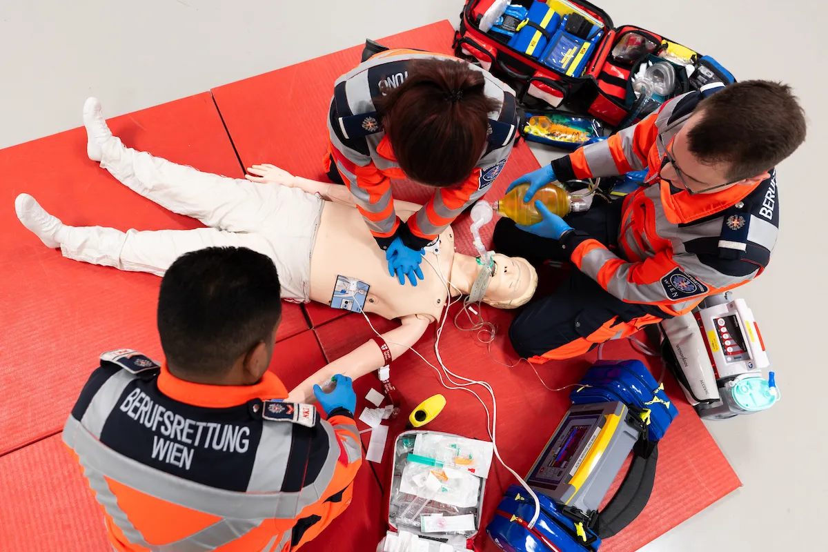 An overhead view of a Berufsrettung Wien emergency medical team in high-visibility uniforms performing advanced life support and CPR on a patient manikin during a realistic first responder training simulation.