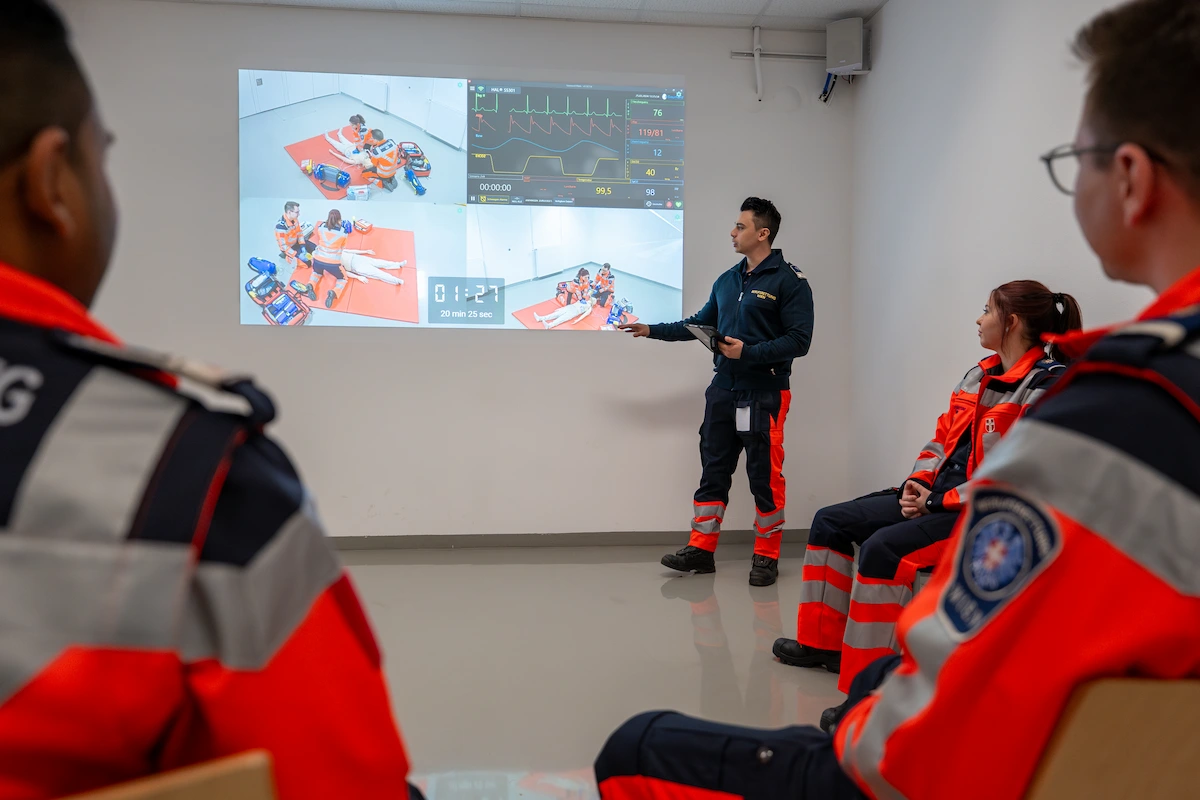 Professional rescue workers from Vienna attending medical simulation briefing with projected vital signs and scenario data.