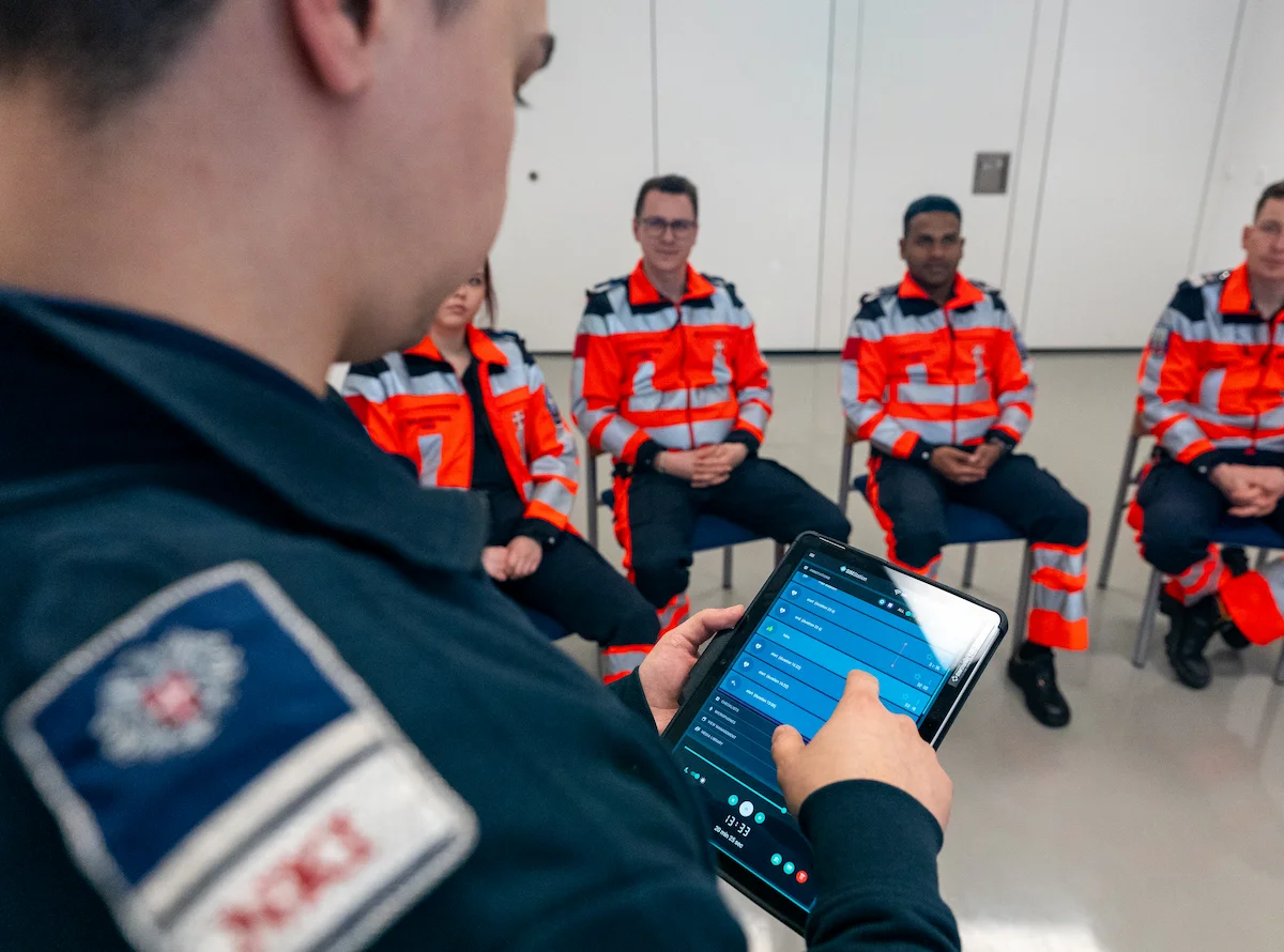 Emergency medical team in orange uniforms receiving digital briefing with tablet during simulation training session.
