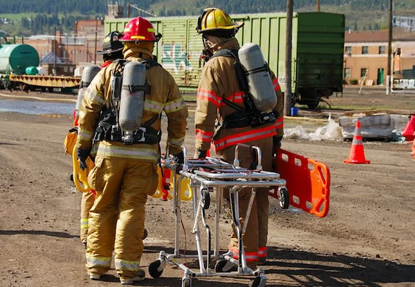 Firefighters in protective gear with oxygen tanks preparing stretcher during emergency response near train yard.