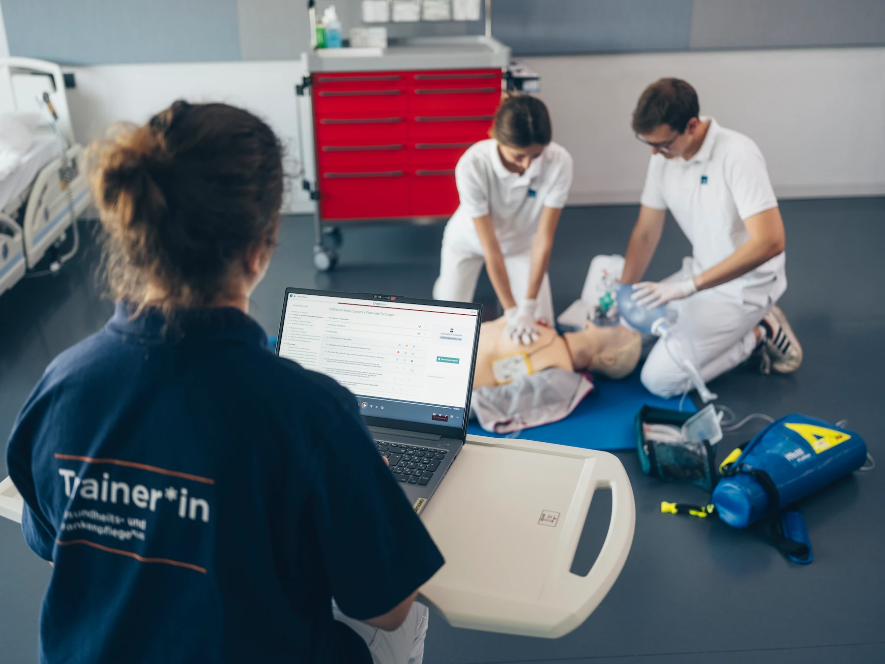 Medical trainees practicing CPR on mannequin with chest compressions and ventilation, guided by trainer using laptop evaluation system.
