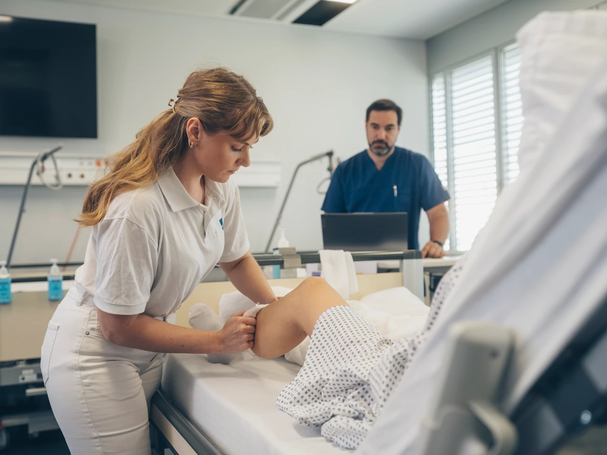Hospital simulation training with healthcare professionals performing patient rehabilitation and documenting care using digital tools.