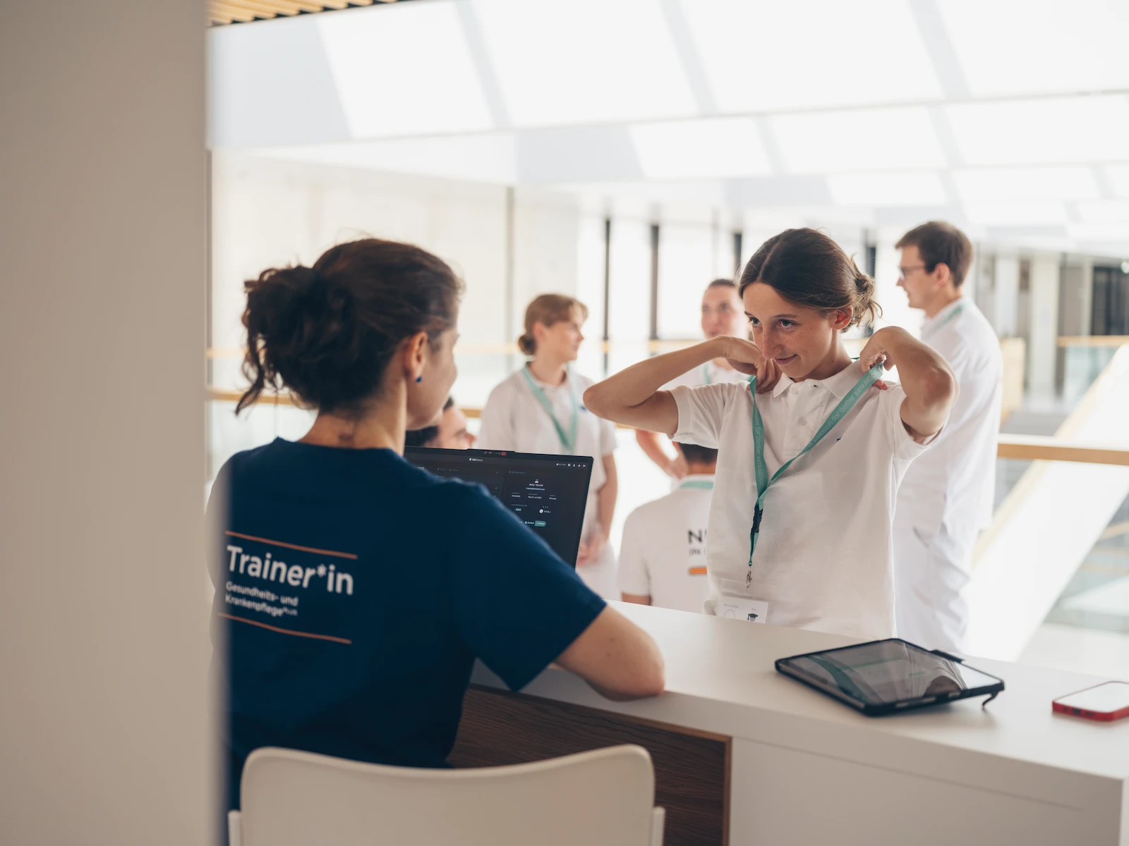 Trainer in nursing education leading healthcare workshop with laptop and tablet, professionals in white uniforms engaged in medical training discussion.