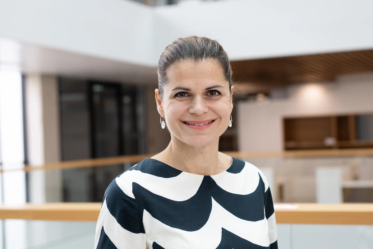Professional portrait of individual in patterned top with earrings standing in modern corporate building with glass panels and wooden accents.