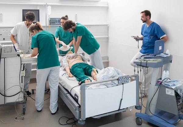 Medical team in scrubs performing emergency simulation with defibrillator on mannequin in hospital training room.