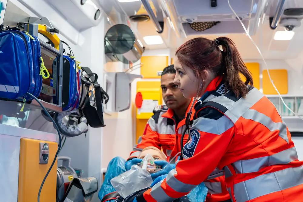 Emergency medical responders in orange uniforms practicing procedures on mannequin inside ambulance with medical equipment.
