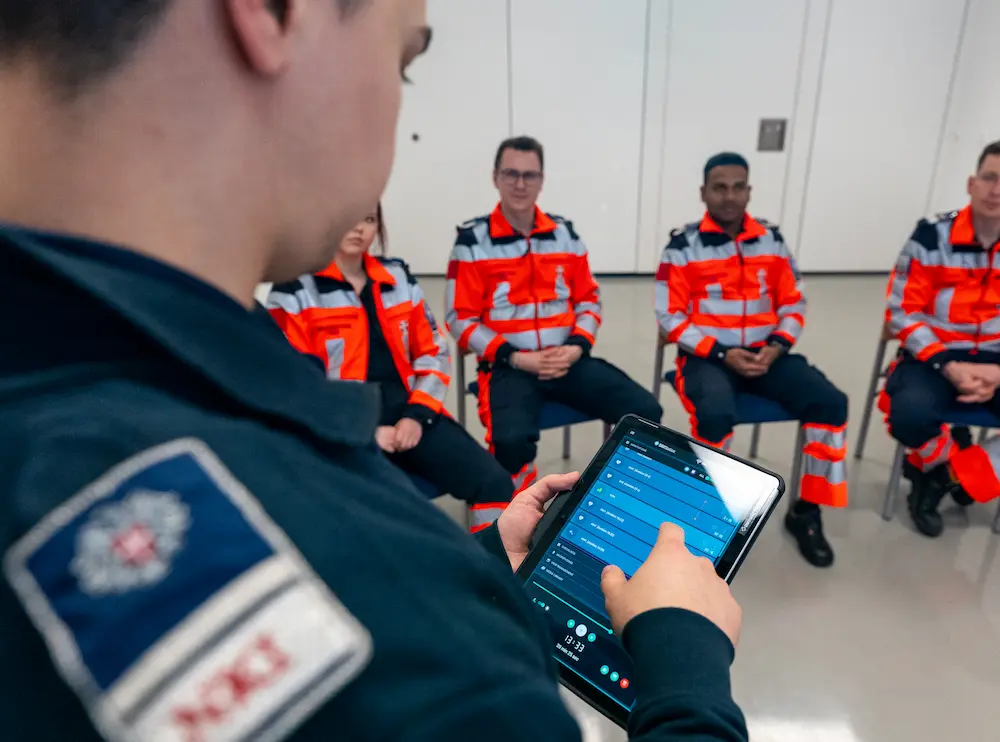 Emergency medical team in orange uniforms receiving digital briefing with tablet during simulation training session.