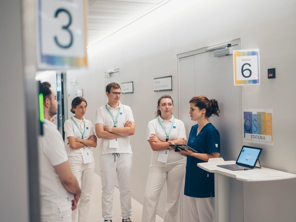 Group of nursing students in white uniforms receiving instructions at OSCE Station 6 during structured clinical skills assessment.