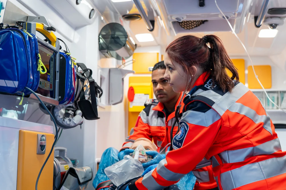 Emergency medical responders in orange uniforms practicing procedures on mannequin inside ambulance with medical equipment.