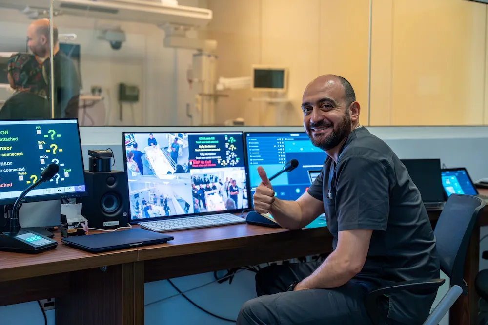 Simulation control room operator smiling with thumbs up, monitoring medical scenarios and patient vitals on multiple screens.
