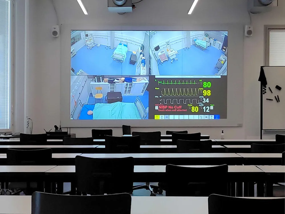 Medical trainees in classroom viewing multi-angle simulation feeds with vital signs monitor for patient care training.