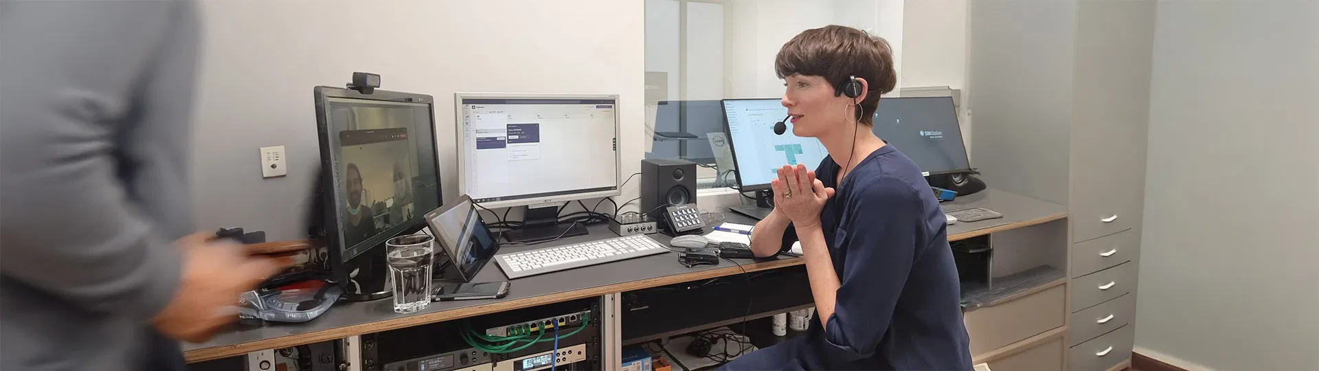 Professional in headset at workstation with multiple monitors engaged in video conference and remote collaboration using communication equipment.