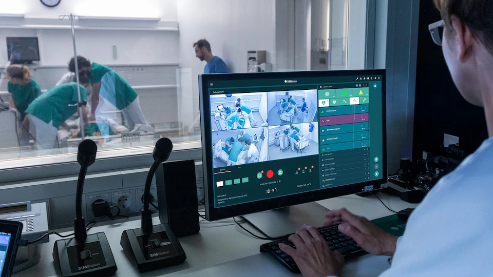 A technician in a simulation control room using SIMStation software to record and monitor a multi-camera medical training session through a one-way mirror.