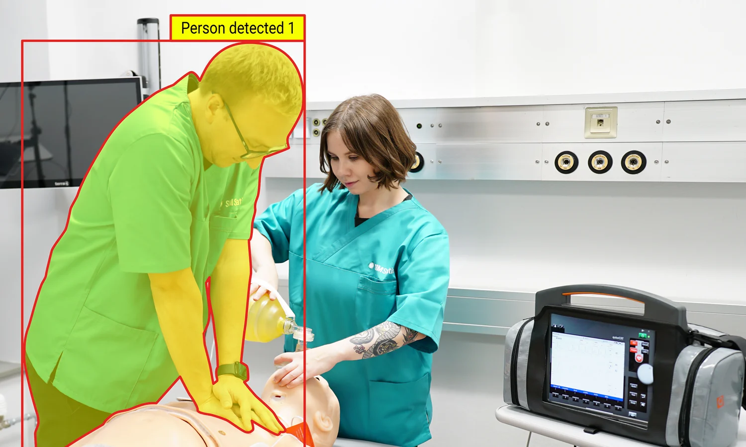 A person in a green uniform practices a medical procedure on a mannequin, assisted by someone in teal scrubs.