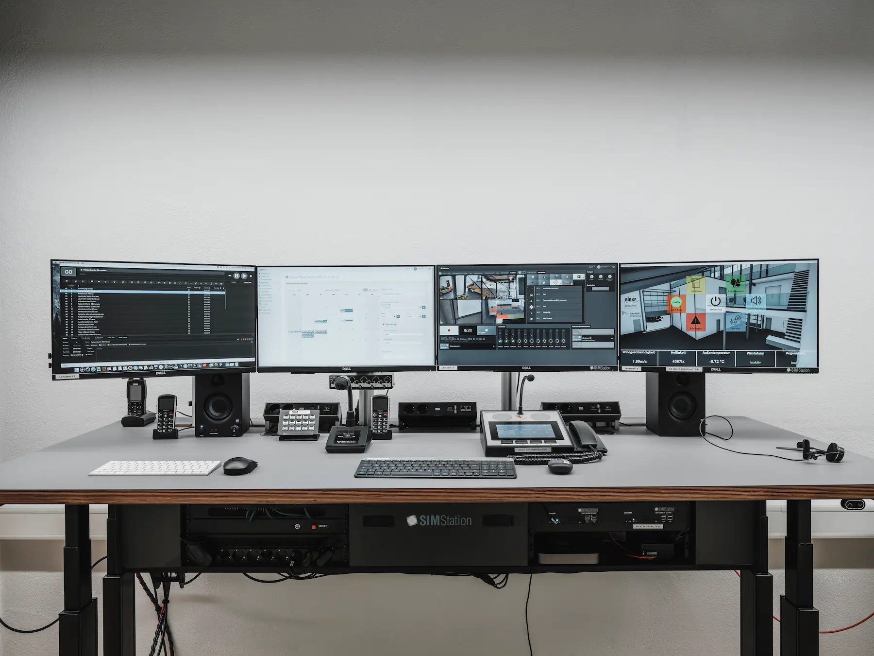 A professional SIMStation simulation control desk featuring a four-monitor setup for recording, scheduling, and live AV monitoring, integrated with high-end audio hardware and rack-mounted processing units.