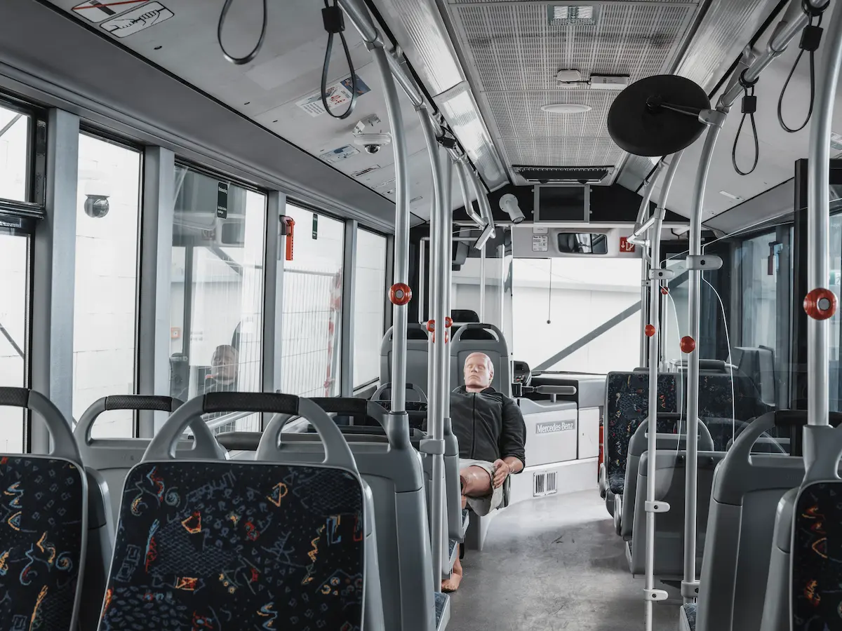 Interior of a modern bus with patterned seats and a life-size mannequin seated mid-vehicle, resembling a training setup.