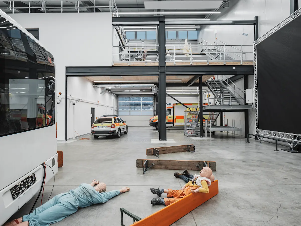 A training facility interior with emergency vehicles, two medical mannequins on the floor, and industrial equipment in a spacious, well-lit area.