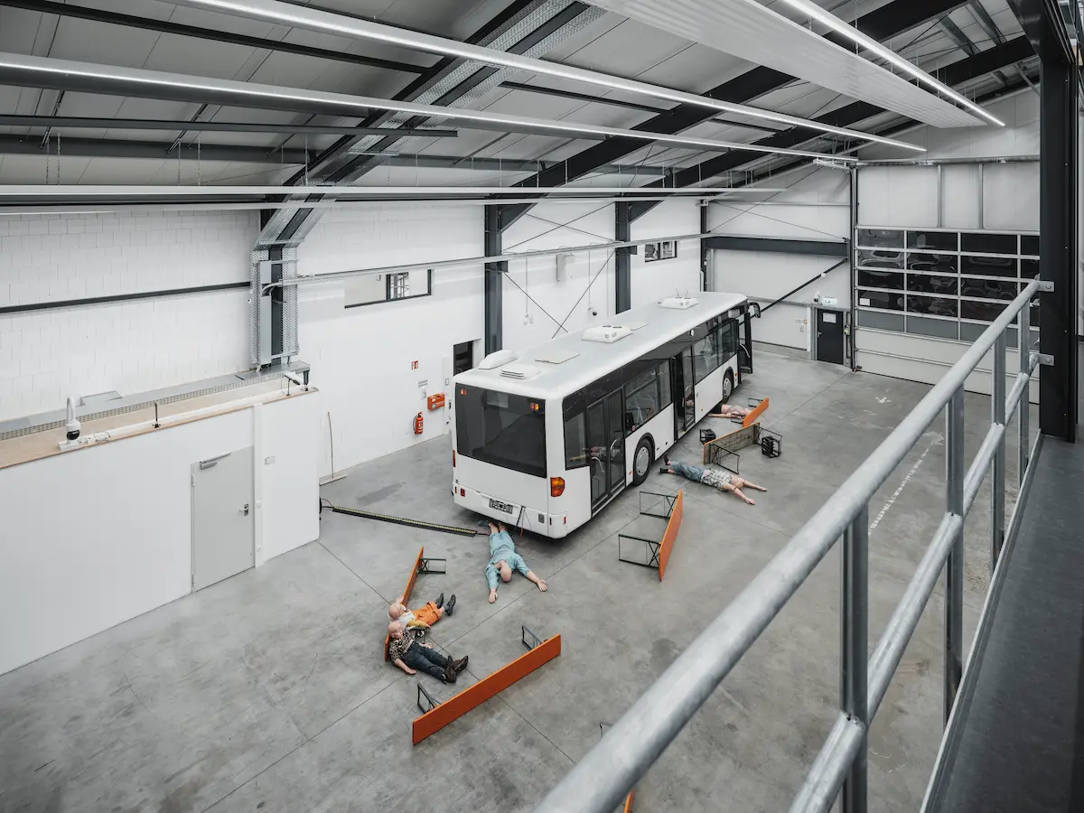 A wide-angle overhead view of a disaster simulation in a training hangar, showing a bus surrounded by simulated casualties for evaluating large-scale team coordination.