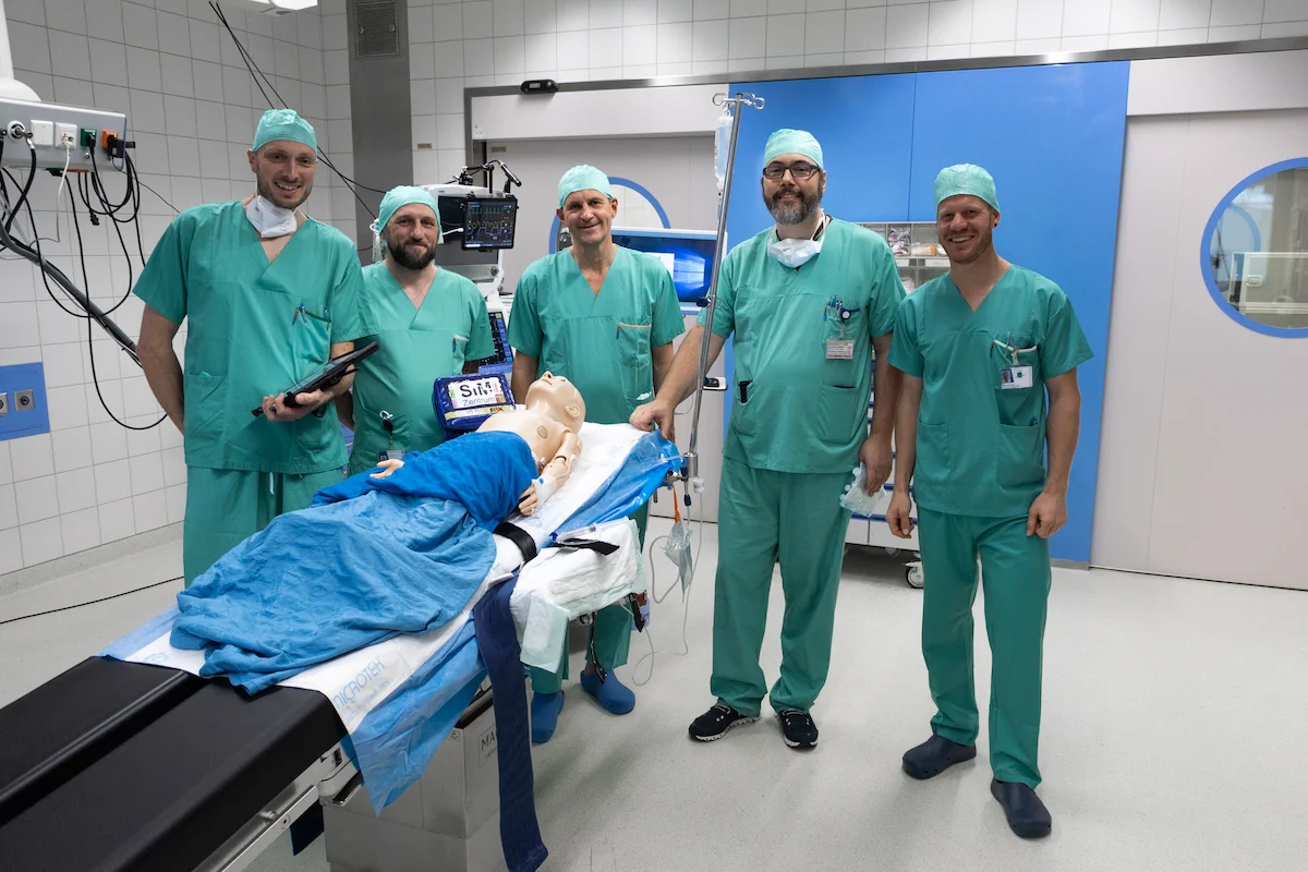Medical team in surgical scrubs conducting simulation training on mannequin in operating room with clinical equipment.