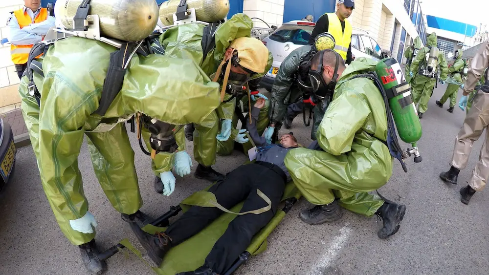 Emergency responders in hazmat suits and gas masks evacuating patient on stretcher during hazardous materials drill in urban setting.