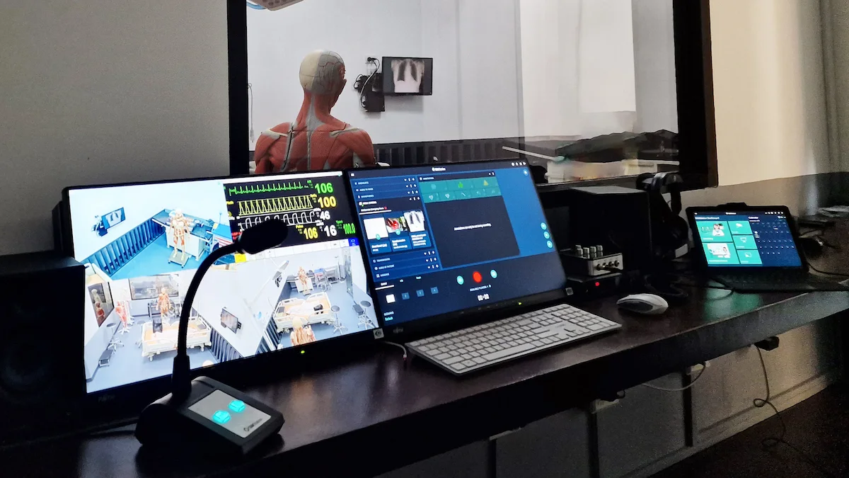 A control room with multiple monitors displays medical data and video feeds. Beyond a window, a medical mannequin is visible in a simulated clinical space, powered by SIMStation Integrated Systems’ highly configurable AV infrastructure for training environments.