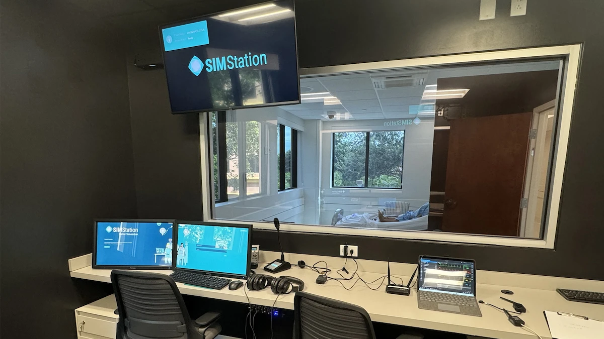 A control room with multiple monitors, computers, headsets, and a large display screen shows the SIMStation logo. The highly configurable AV infrastructure supports complex, multi-room scenarios and unifies recording for demanding simulation environments.