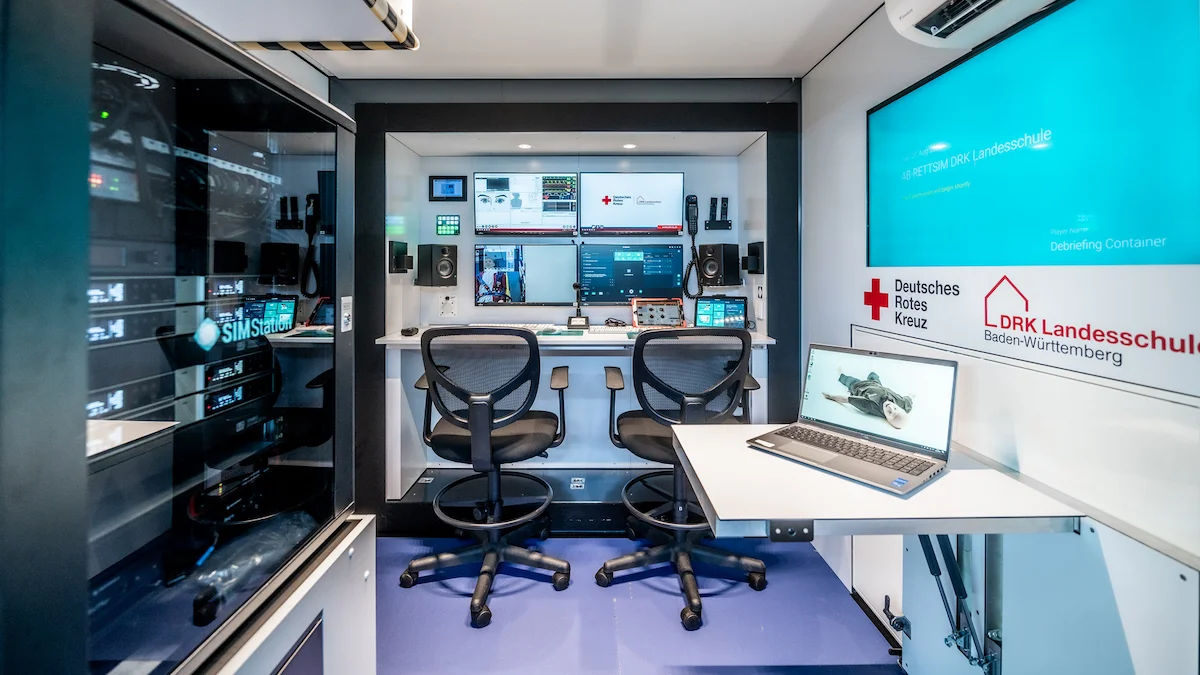 Interior of a modern mobile command center with two chairs, multiple computer monitors, server-based architecture, and a laptop on a side desk—enabling live control and analysis within one coordinated setup. Walls display Deutsches Rotes Kreuz and DRK Landesschule logos.