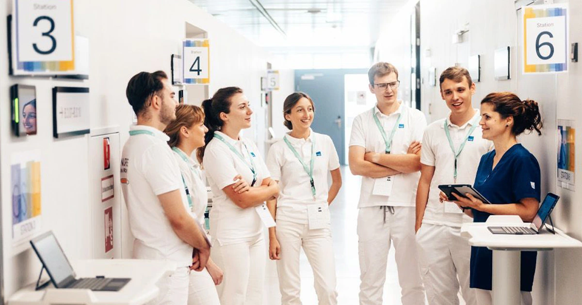 A group of medical students in white uniforms and lanyards standing in a bright corridor, engaging in a clinical briefing or debriefing session with an instructor using a tablet.