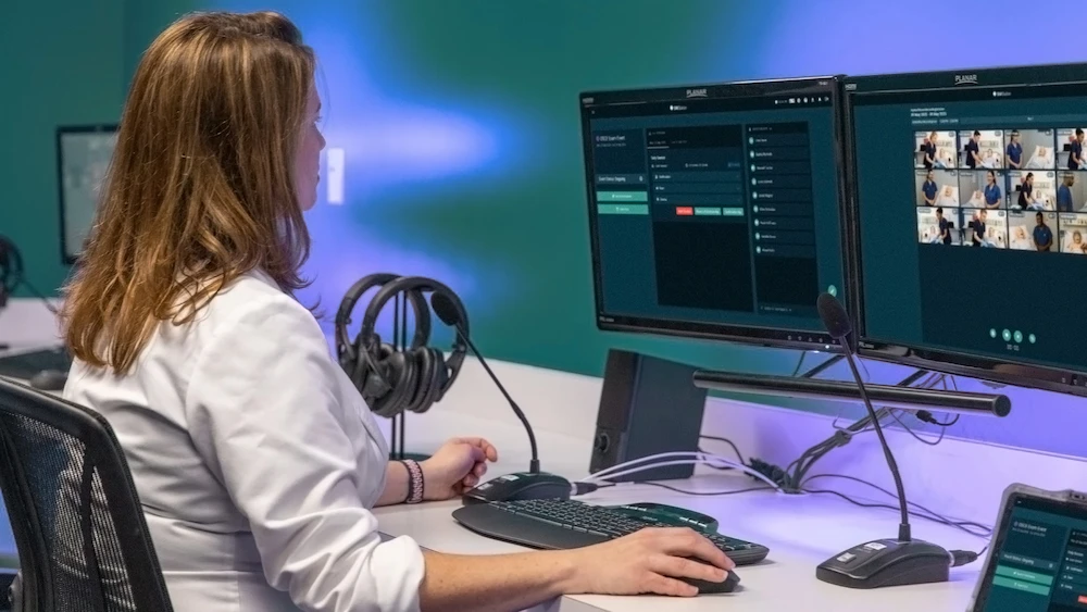 Medical professional in white coat monitoring hospital scenarios on dual screens with surveillance feeds and control panel.