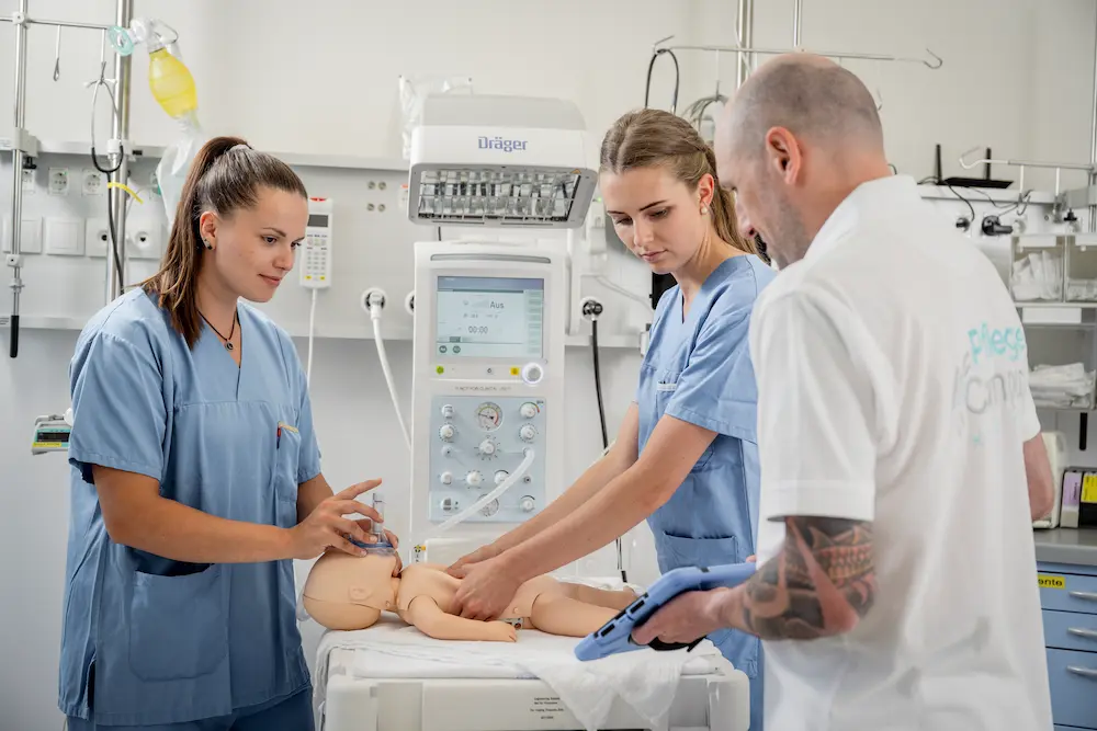 Medical trainees in blue scrubs practice neonatal resuscitation on an infant manikin under a Dräger warming station while an instructor monitors the session using a digital tablet.