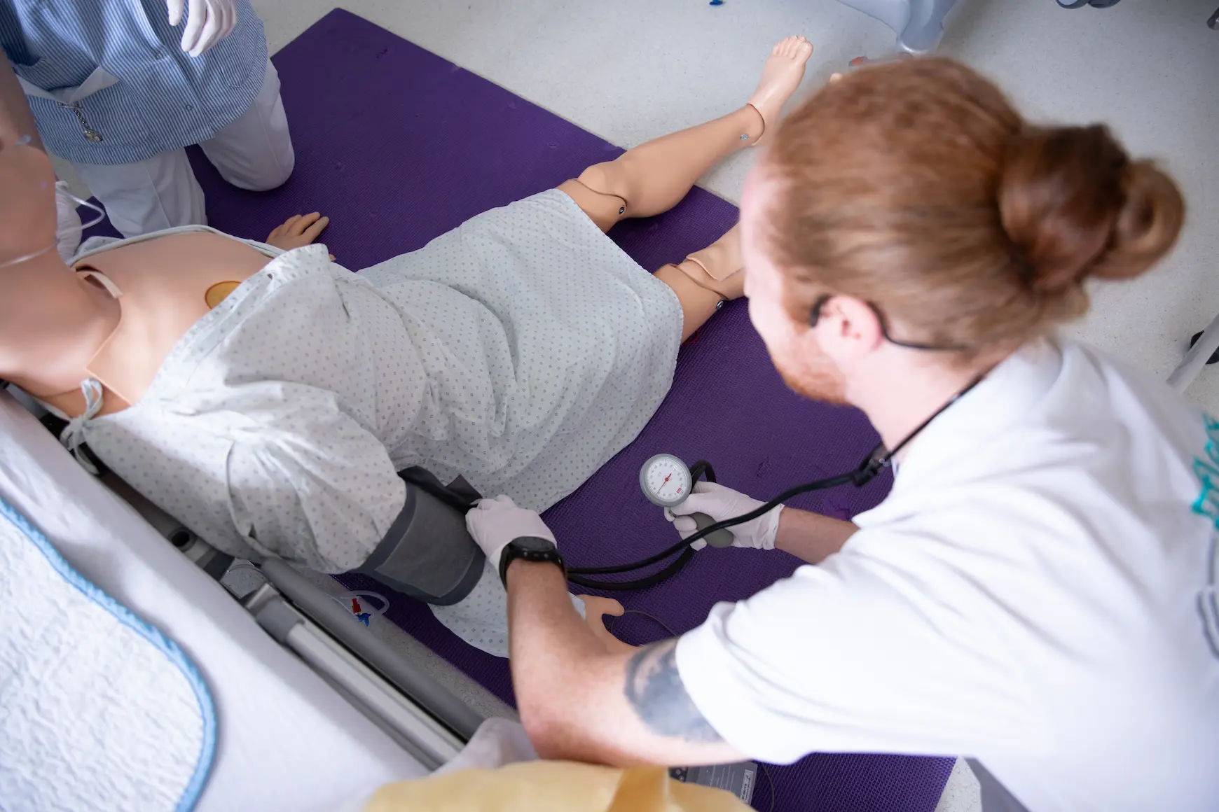 A medical student practicing manual blood pressure measurement on a patient manikin lying on a purple mat during a clinical skills training session.