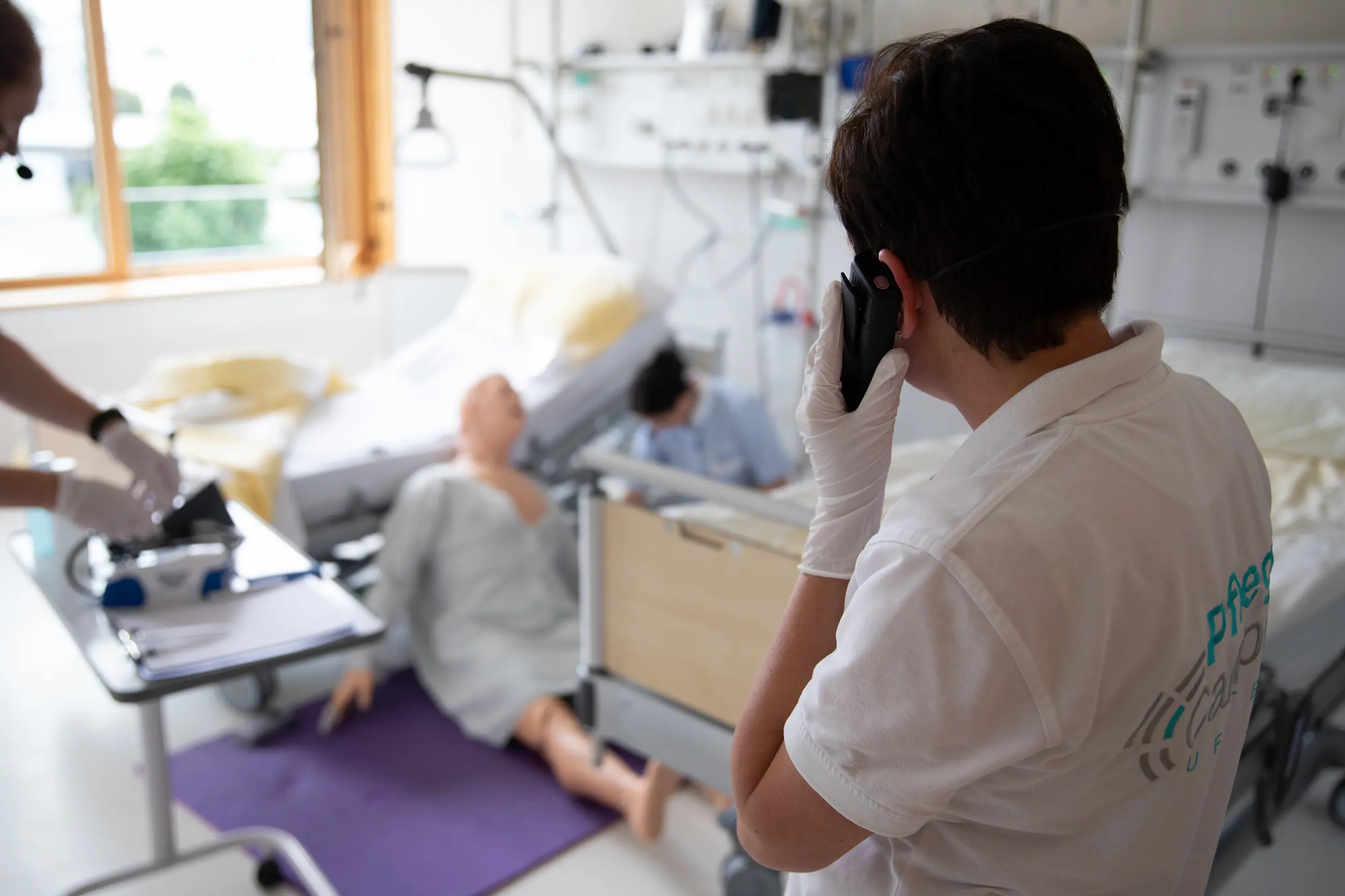 A medical trainee in a white uniform and gloves talks on a mobile phone during a high-pressure simulation, coordinating emergency response for a patient manikin in a hospital ward setting.