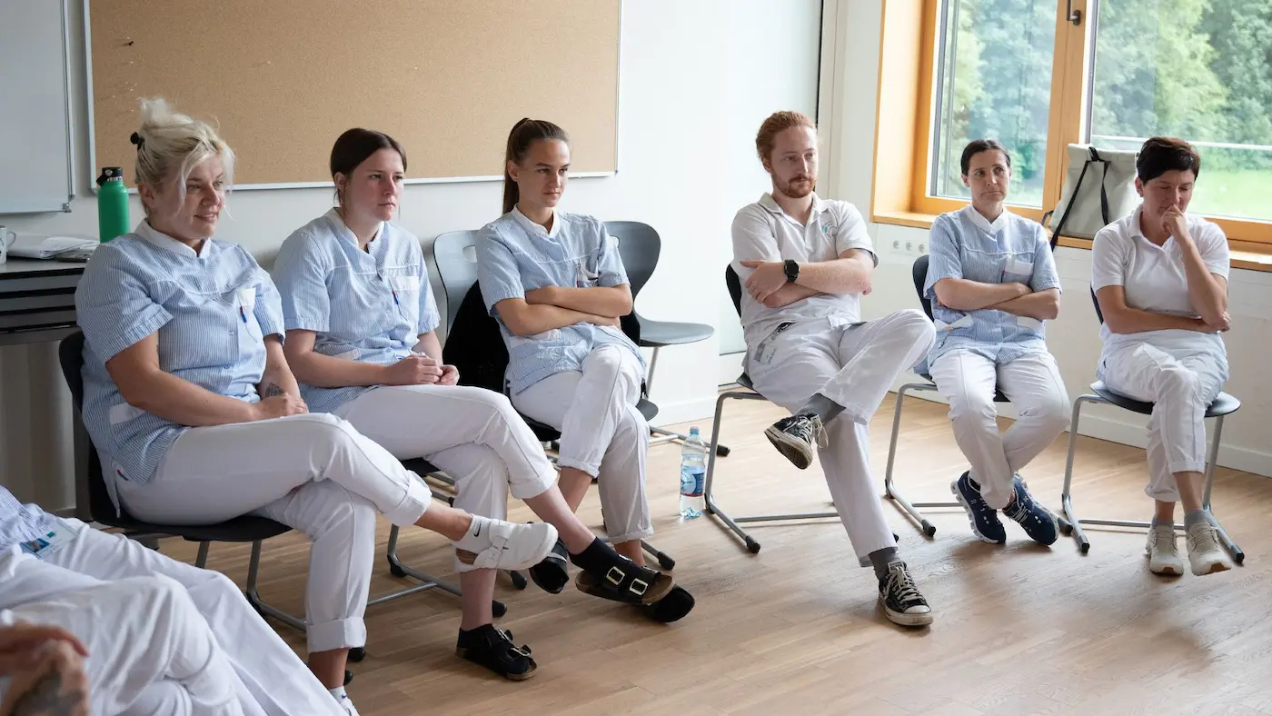 A group of medical trainees in nursing uniforms sitting in a circle during a reflective debriefing session after a clinical simulation at a healthcare training center.