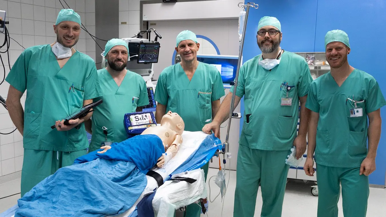 Medical team in surgical scrubs conducting simulation training on mannequin in operating room with clinical equipment.