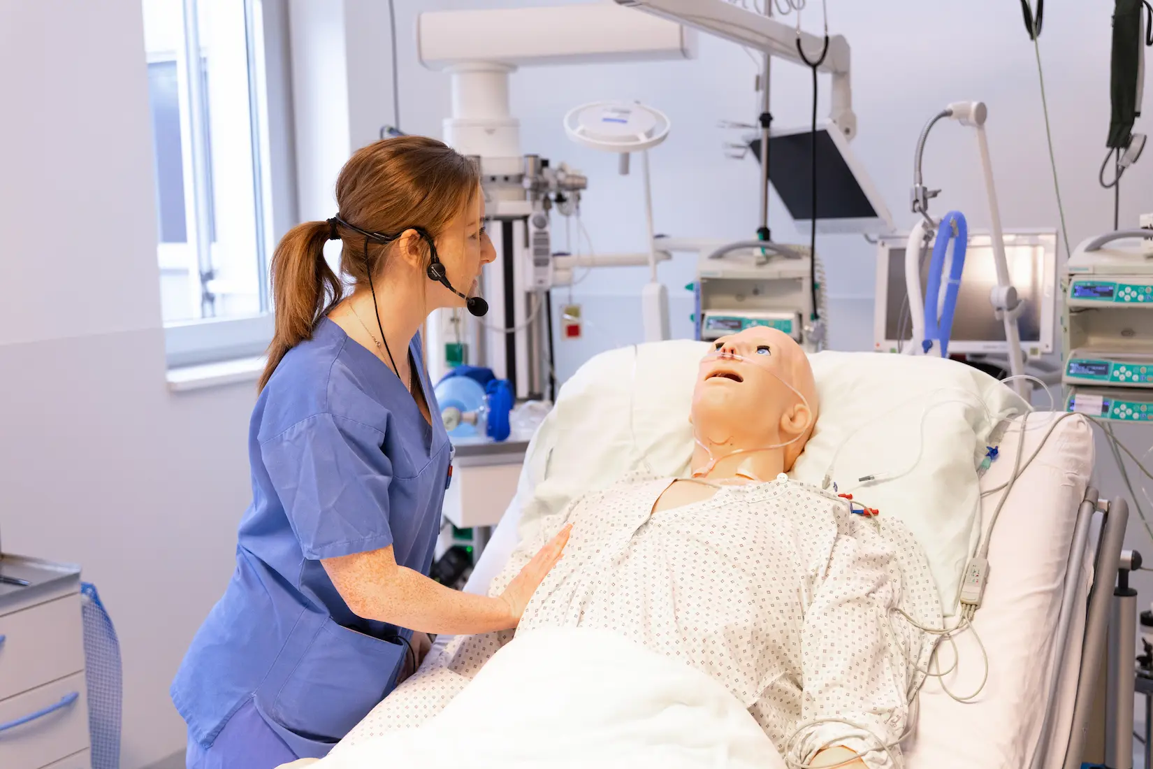 A medical instructor wearing a headset microphone monitors a high-fidelity patient manikin in a simulated hospital ward, providing real-time guidance during a clinical training session.