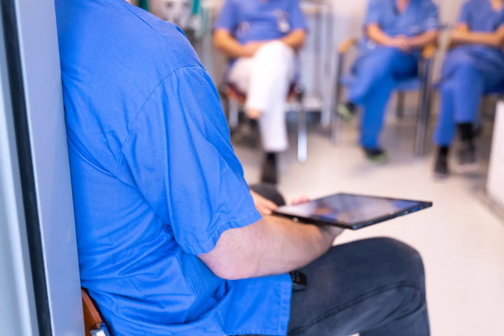 Over-the-shoulder view of a medical instructor in blue scrubs holding a digital tablet during a collaborative debriefing session with a group of healthcare trainees.