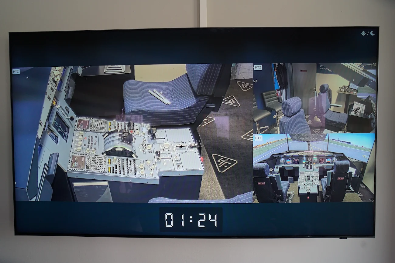 A large wall-mounted monitor displaying a SIMStation multi-view interface of an aircraft cockpit simulation, including synchronized views of the center console, pilot seating, and the primary flight display with a digital timestamp.
