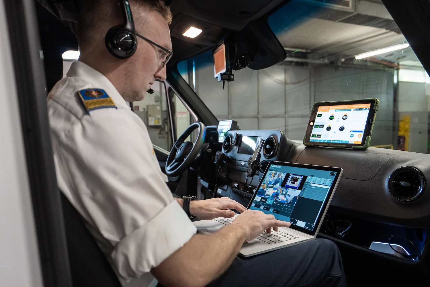A man in uniform sits in a vehicle equipped with SIMStation Mobile Systems, wearing a headset and typing on a laptop.