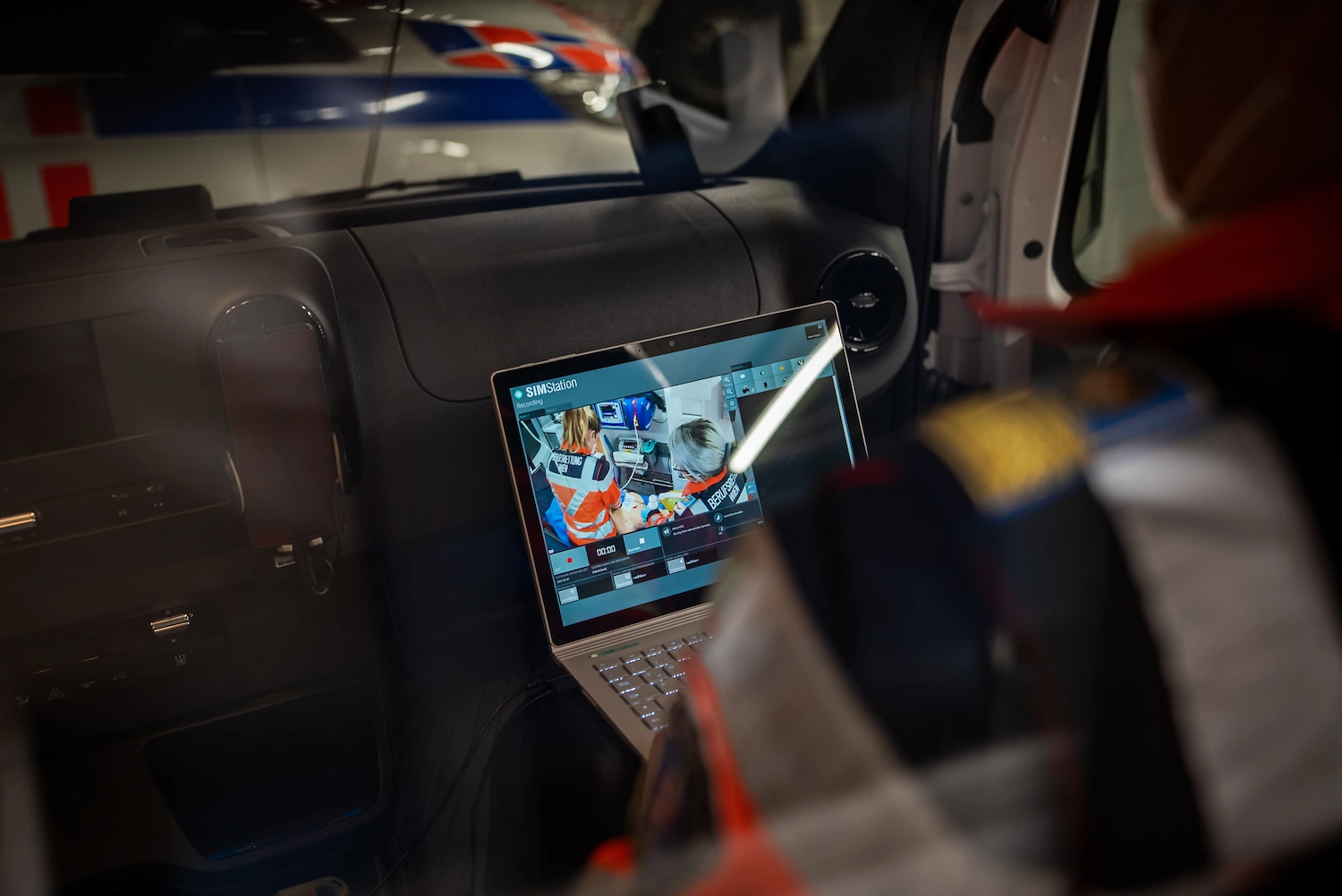 A person sits inside a vehicle equipped with SIMStation Mobile Systems, looking at a laptop screen showing a video call with several people.