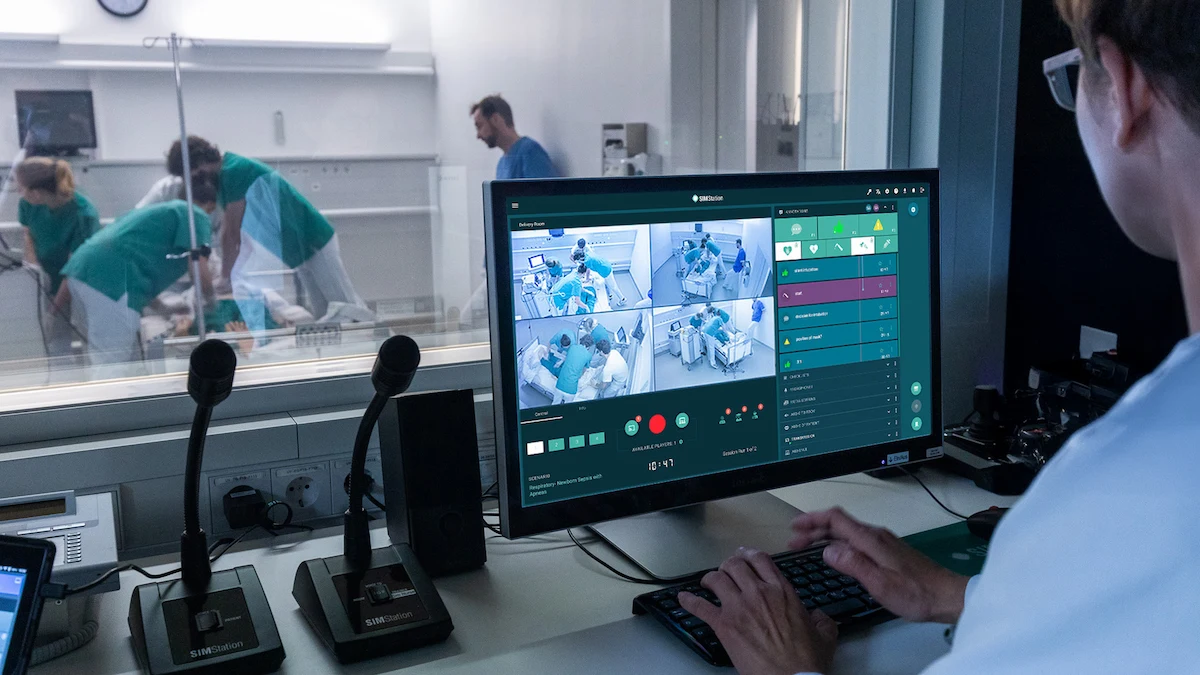 A person monitors hospital room camera feeds on a computer screen from a control room, ensuring every phase of your simulation workflow comes together seamlessly while medical staff attend to a patient in an adjacent room.